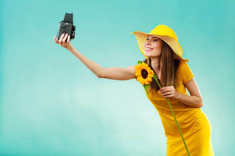 Summer woman wearing yellow dress and hat with sunflower taking self picture with old vintage camera on vivid blue background